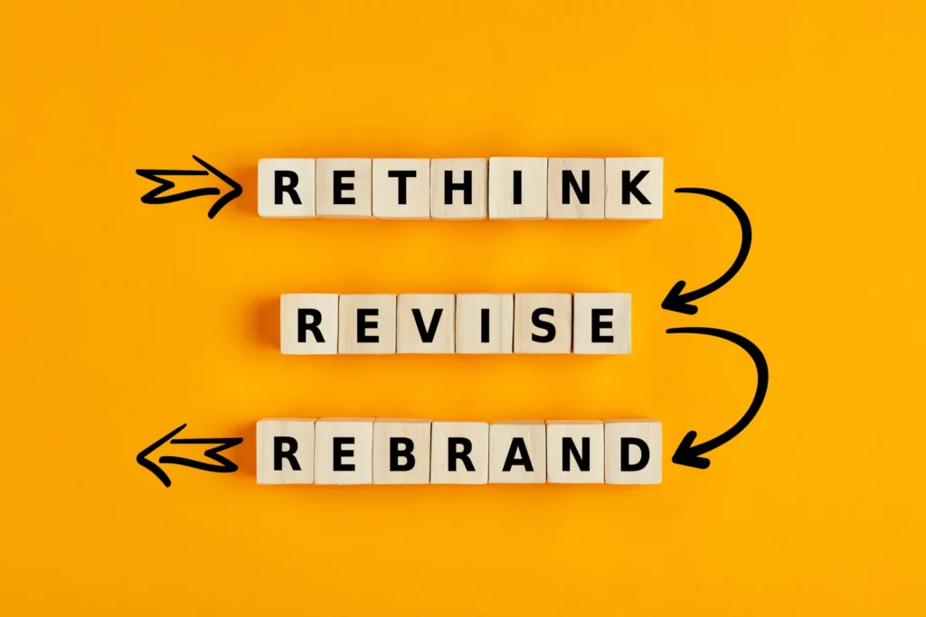 Blocs de bois alignés avec les mots "Rethink", "Revise", "Rebrand" sur fond jaune, illustrant les étapes du rebranding.