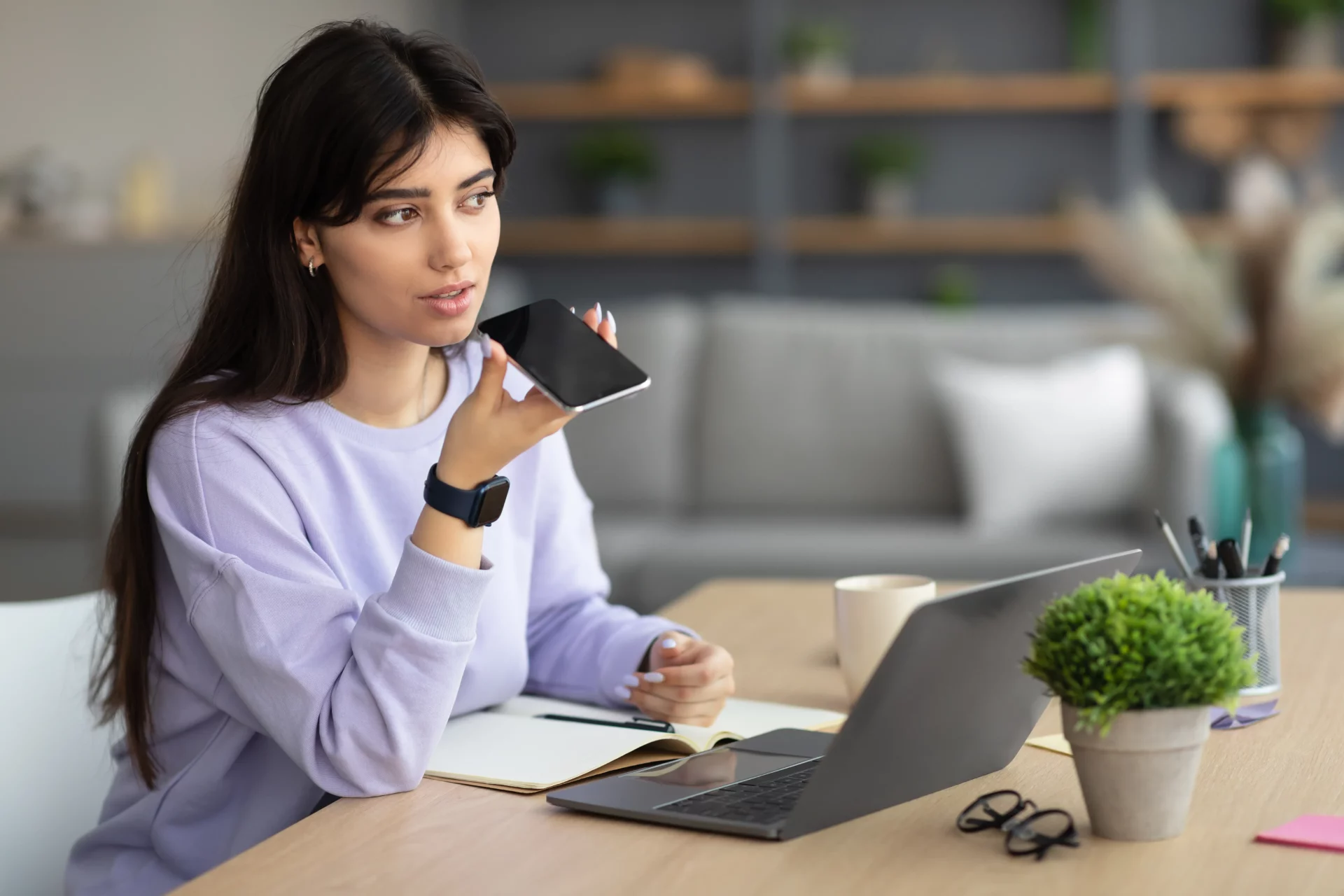 Une femme assise à un bureau, tenant un smartphone et enregistrant un message vocal, avec un ordinateur portable, un carnet, et des accessoires de bureau en arrière-plan.