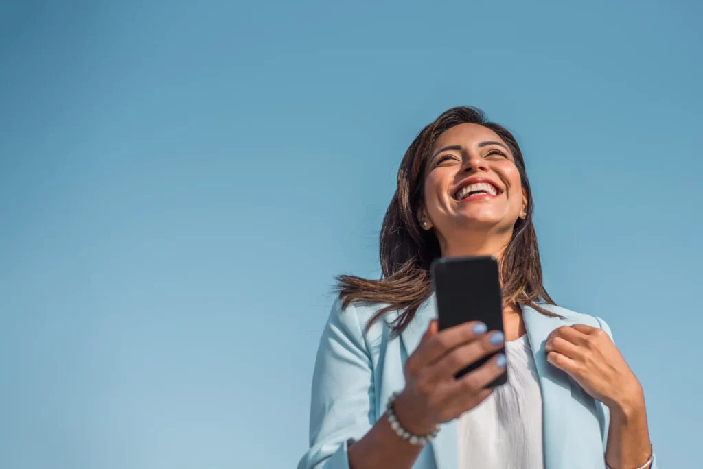 Femme tenant un smartphone et souriant, avec un ciel bleu en arrière-plan