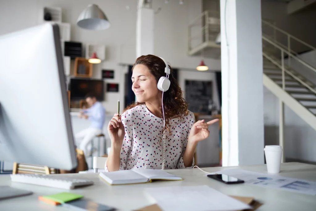 Femme portant des écouteurs blancs et écoutant de la musique tout en travaillant à son bureau dans un espace de travail moderne.