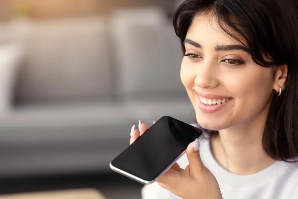 Femme souriante enregistrant un message vocal sur son téléphone, illustrant l'importance de l'audio dans la communication interne.